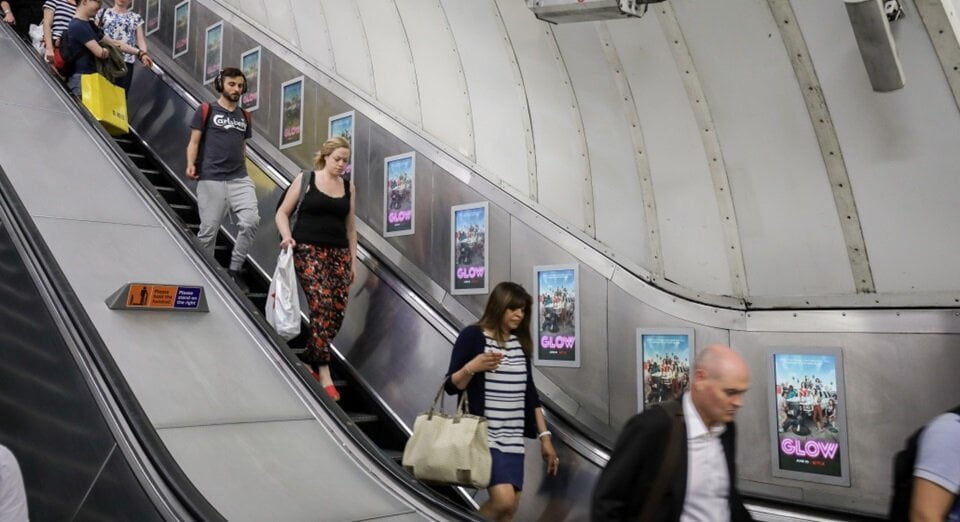 People going down the escalator towards the London Underground with digital escalator panels advertising to them.