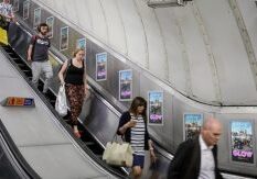 People going down the escalator towards the London Underground with digital escalator panels advertising to them.
